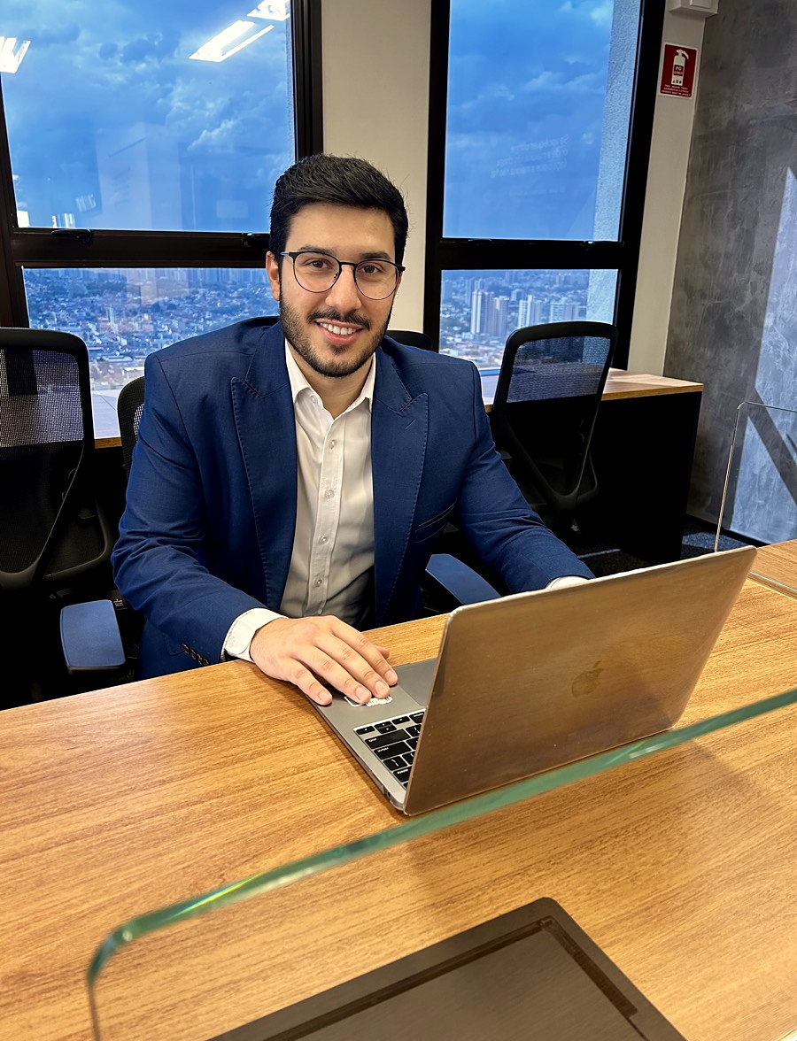 Bruno de Oliveira sentado à mesa de reunião do escritório, notebook aberto e a mão no teclado, olhando para quem está do outro lado da mesa