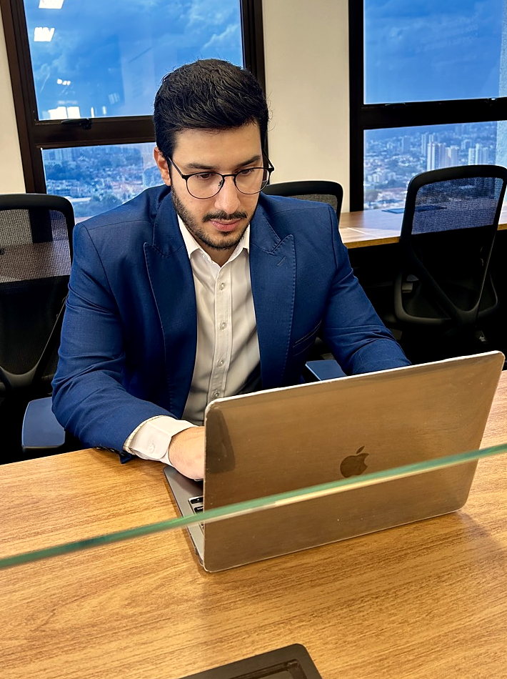 Bruno de Oliveira de cabeça baixa diante do notebook, lendo os números de um mês, com a mesa de madeira do escritório em primeiro plano